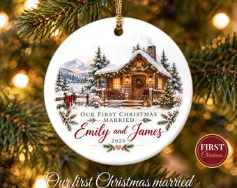 May include: A round white ceramic Christmas ornament featuring a winter cabin scene with snow-covered mountains and trees. The text reads: OUR FIRST CHRISTMAS MARRIED, Emily and James, 2026. It hangs from a gold cord.