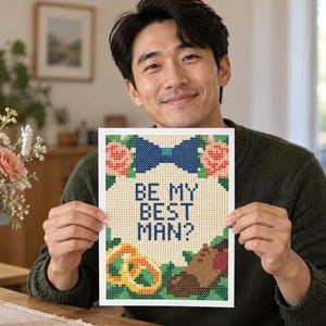 May include: A rectangular cross-stitch pattern on white fabric featuring the text BE MY BEST MAN? in blue, surrounded by a blue bow tie, pink flowers, two gold rings, and a brown dress shoe, all rendered in a pixelated grid design style.