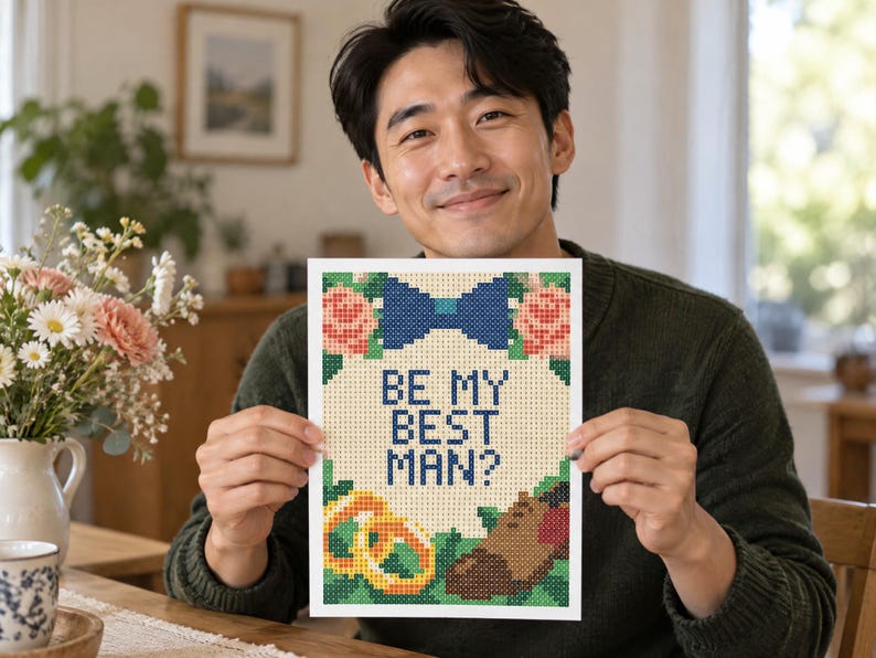 May include: A rectangular cross-stitch pattern on white fabric featuring the text BE MY BEST MAN? in blue, surrounded by a blue bow tie, pink flowers, two gold rings, and a brown dress shoe, all rendered in a pixelated grid design style.