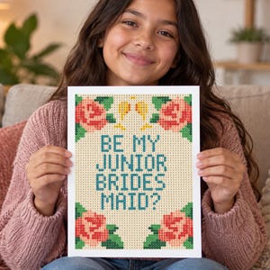 May include: A rectangular print featuring a pixelated design on a light beige background. The text reads BE MY JUNIOR BRIDES MAID? in teal letters. The design is framed by four clusters of pink roses with green leaves and two small yellow glasses.