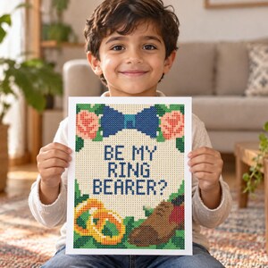 May include: A rectangular cross-stitch pattern on white fabric featuring the text BE MY RING BEARER? in dark blue. The design includes a blue bow tie at the top, two gold rings on the bottom left, and a brown shoe on the bottom right with roses.