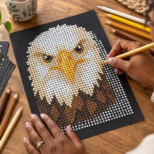 May include: A black paper sheet featuring a dot-to-dot or grid-based illustration of a bald eagle head with a yellow beak, white head feathers, and brown neck feathers, being colored with a yellow pencil on a wooden surface.