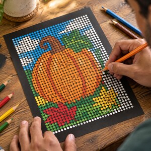 May include: A dot-to-dot coloring page on black paper featuring a large orange pumpkin, green leaves, and a red maple leaf. The design is composed of a grid of white circles waiting to be filled with color, with a pencil currently coloring a circle.