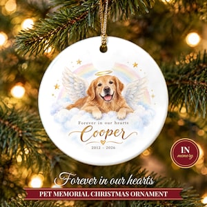 May include: A round white ceramic Christmas ornament featuring a golden retriever with angel wings and a halo, resting on clouds under a rainbow. The text reads Forever in our hearts, Cooper, and the years 2012-2026 with a small heart.