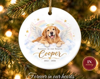 May include: A round white ceramic Christmas ornament featuring a golden retriever with angel wings and a halo, resting on clouds under a rainbow. The text reads Forever in our hearts, Cooper, and the years 2012-2026 with a small heart.