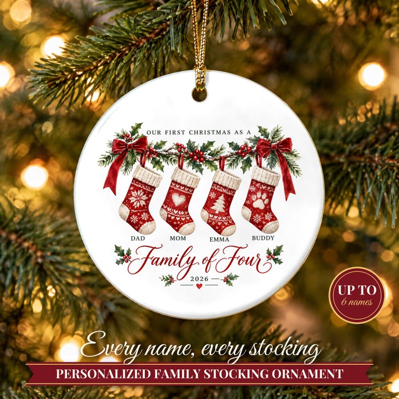 May include: A round white ceramic Christmas ornament featuring four red and white patterned stockings hanging from a green holly garland with red bows. Text reads: OUR FIRST CHRISTMAS AS A Family of Four 2026, with names DAD, MOM, EMMA, BUDDY.