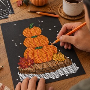 May include: A diamond painting kit on black paper featuring a stack of three orange pumpkins, a red maple leaf, and a yellow leaf on a brown wooden surface. The design includes white circular accents and a grid of letters for color placement.