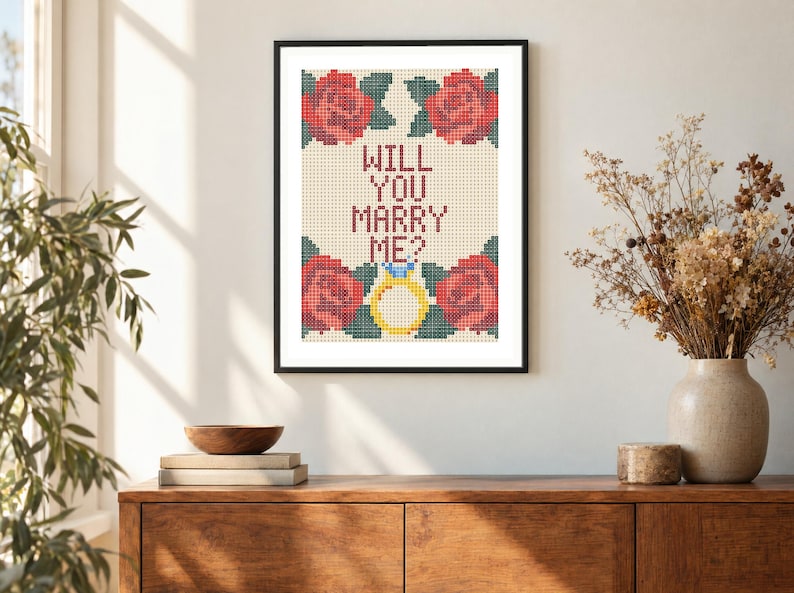 May include: A framed cross-stitch embroidery featuring the text WILL YOU MARRY ME? in dark red letters, surrounded by four red roses with green leaves and a yellow engagement ring with a blue gemstone, all set against a cream-colored grid.