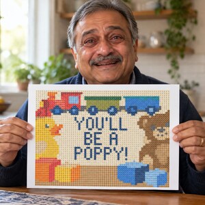Puede incluir: Una impresi&oacute;n rectangular con un dise&ntilde;o pixelado y el texto YOU'LL BE A POPPY! en azul oscuro. La imagen incluye un tren rojo y azul, un pato de goma amarillo, un oso de peluche marr&oacute;n y bloques azules sobre un fondo de color beige.