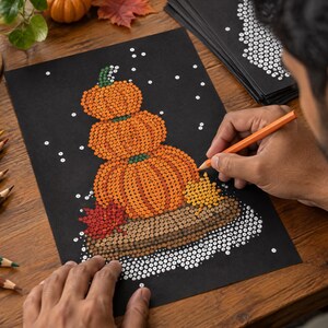 May include: A black sheet of paper featuring a dot art illustration of three stacked orange pumpkins on a brown log, accented with a red maple leaf and yellow foliage, surrounded by white dots representing snow on a dark background.