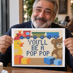 May include: A rectangular cross-stitch pattern featuring a red train, a yellow duck, a brown teddy bear, and blue blocks. The text in the center reads YOU'LL BE A POP POP in dark blue letters against a light tan background with a grid design.