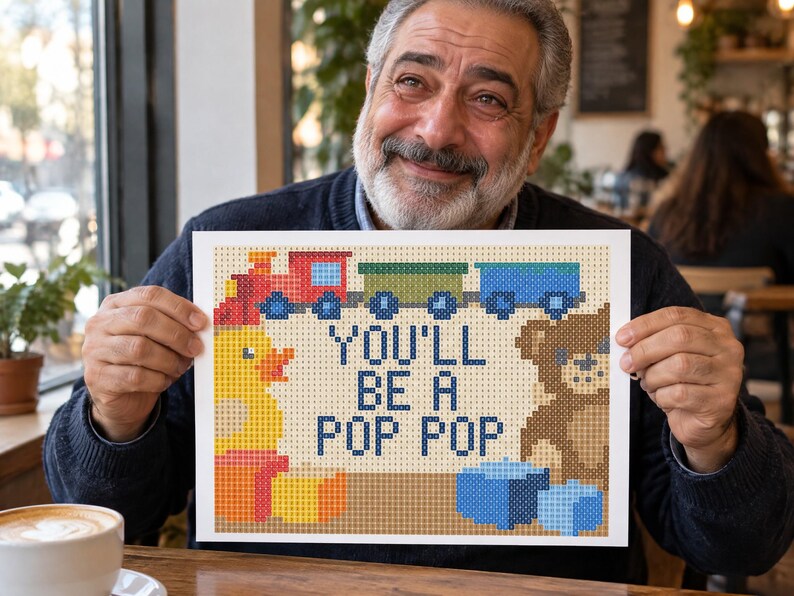 May include: A rectangular cross-stitch pattern featuring a red train, a yellow duck, a brown teddy bear, and blue blocks. The text in the center reads YOU'LL BE A POP POP in dark blue letters against a light tan background with a grid design.