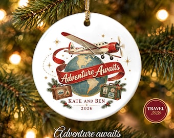 May include: A round white ceramic ornament featuring a red and white airplane flying over a globe. A red ribbon banner reads Adventure Awaits. Below are two vintage suitcases, the text Kate and Ben, and the year 2026 with pine sprigs.