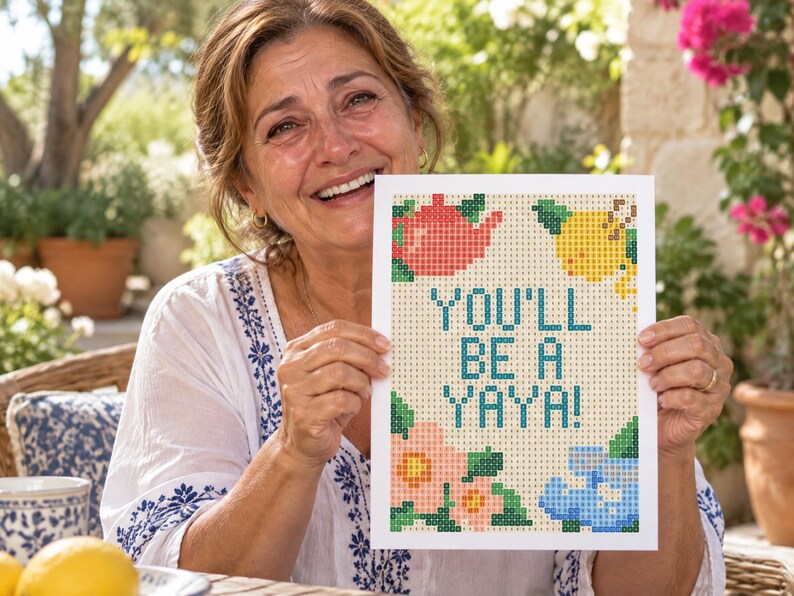 K&ouml;nnte beinhalten: Eine rechteckige wei&szlig;e Karte mit einem pixeligen Kreuzstich-Design und dem Text "YOU'LL BE A YAYA!" in Blau. Das Motiv zeigt eine rote Teekanne, eine gelbe Zitrone, eine rosa Blume und blaue Blumen, die alle in einem Rastermuster dargestellt sind.