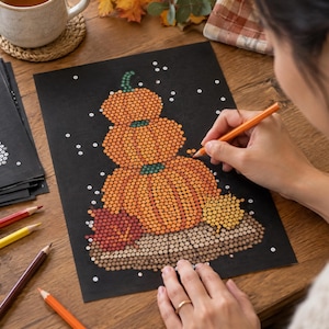 May include: A black sheet of paper featuring a dot-art illustration of three stacked orange pumpkins with green stems, surrounded by autumn leaves. The artwork is created using small circular colored dots, with an orange pencil held to add detail.