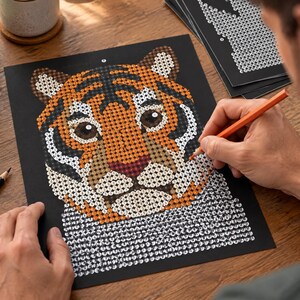 May include: A paint-by-number activity sheet featuring a tiger head illustration on a black background. The tiger is composed of small numbered circles in shades of orange, black, white, and brown. An orange pencil is used to fill in the design.