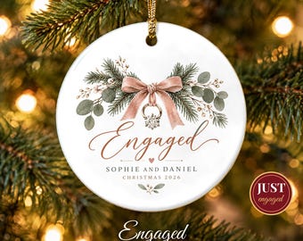 May include: A round white ceramic ornament featuring a pink ribbon tied around a diamond ring, surrounded by pine branches and eucalyptus leaves. The text reads Engaged, Sophie and Daniel, and Christmas 2026, with a gold hanging cord.