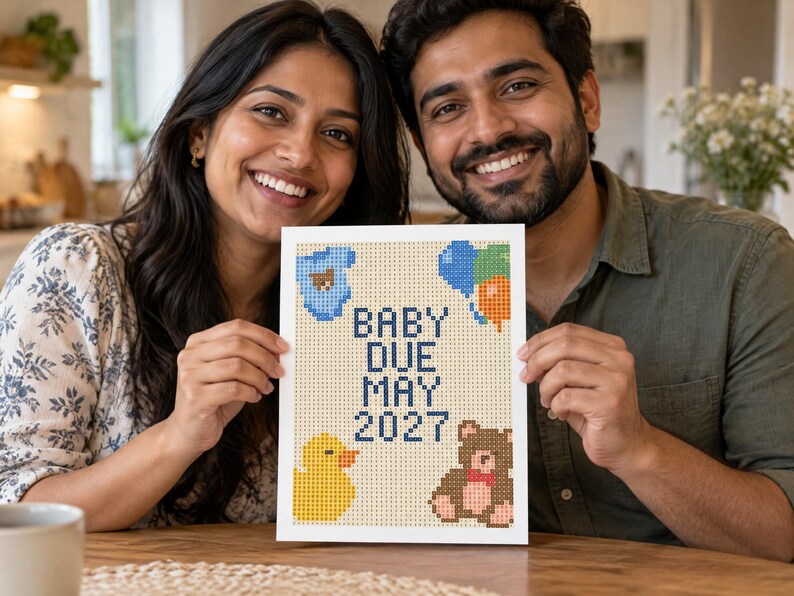 May include: A rectangular white sign featuring a pixelated cross-stitch design with the text BABY DUE MAY 2027 in dark blue. The design includes a blue baby onesie, a cluster of blue and orange balloons, a yellow rubber duck, and a brown teddy bear.