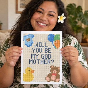 May include: A rectangular white card featuring a pixelated cross-stitch design with the text WILL YOU BE MY GOD MOTHER? in dark blue. The design includes a light blue baby onesie, a cluster of blue, green, and orange balloons, a yellow duck, and a brown teddy bear.