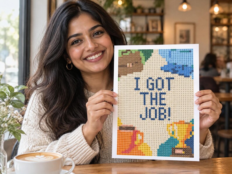 May include: A rectangular diamond painting craft project featuring a grid pattern with the text I GOT THE JOB! in dark blue. The design includes colorful pixelated illustrations of a brown briefcase, a gold trophy, and a coffee mug on a light background.