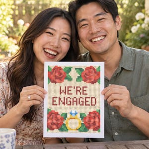 K&ouml;nnte beinhalten: Ein rechteckiges wei&szlig;es Schild mit einem pixeligen Kreuzstich-Design und dem Text WE'RE ENGAGED in Dunkelrot. Das Motiv zeigt vier rote Rosen mit gr&uuml;nen Bl&auml;ttern in den Ecken sowie einen gelben Verlobungsring mit einem blauen Diamanten.