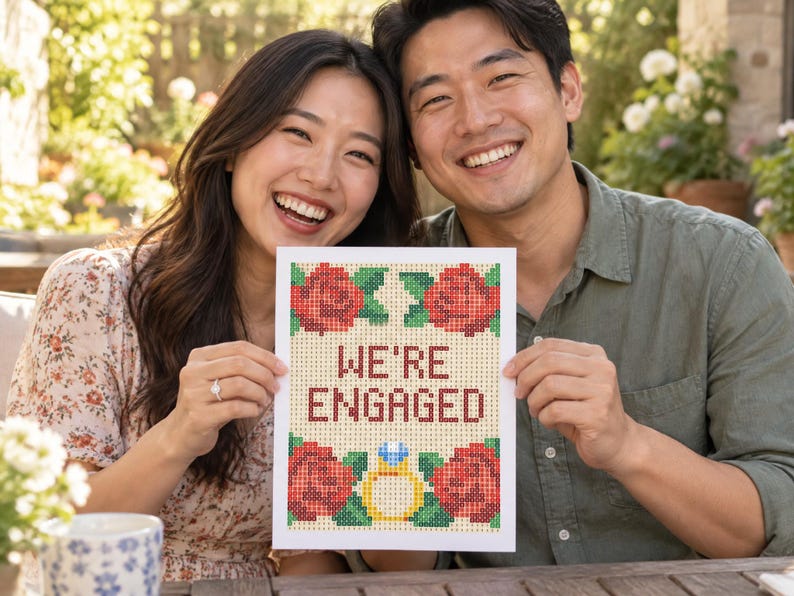 K&ouml;nnte beinhalten: Ein rechteckiges wei&szlig;es Schild mit einem pixeligen Kreuzstich-Design und dem Text WE'RE ENGAGED in Dunkelrot. Das Motiv zeigt vier rote Rosen mit gr&uuml;nen Bl&auml;ttern in den Ecken sowie einen gelben Verlobungsring mit einem blauen Diamanten.