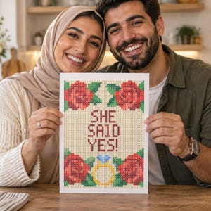 May include: A rectangular print featuring a cross-stitch style design with the text "SHE SAID YES!" in red letters. The design includes four red roses with green leaves in the corners and a yellow engagement ring with a blue diamond at the bottom.