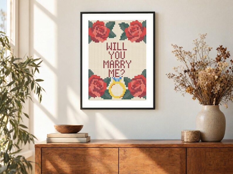 May include: A framed cross-stitch embroidery featuring the text WILL YOU MARRY ME? in dark red letters, surrounded by four red roses with green leaves and a yellow engagement ring with a blue gemstone, all set against a cream-colored grid.