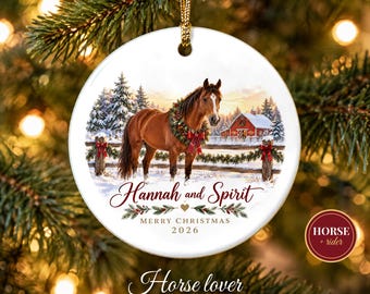 May include: A round white ceramic ornament featuring a brown horse wearing a festive wreath around its neck. The scene includes a snowy landscape, a red barn, and pine trees. Text reads Hannah and Spirit, MERRY CHRISTMAS 2026.