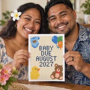 May include: A rectangular white sign featuring a pixelated cross-stitch design with the text BABY DUE AUGUST 2027 in dark blue. The design includes a blue baby onesie, a cluster of blue, green, and orange balloons, a yellow duck, and a brown teddy bear.