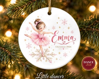 May include: A round white ceramic ornament featuring a illustration of a young ballerina in a pink tutu dancing on a large snowflake. The text reads Emma, Christmas 2026, and is accented with small pink and gold snowflakes and stars.