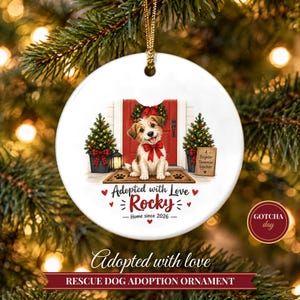 May include: A round white ceramic ornament featuring a brown and white puppy wearing a red bow, sitting on a doormat in front of a red door with two small Christmas trees. The text reads Adopted with Love, Rocky, Home since 2026, and A Brighter Tomorrow Together.