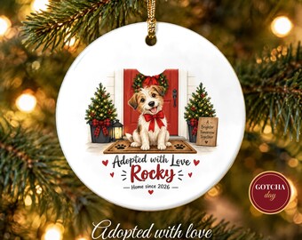 May include: A round white ceramic ornament featuring a brown and white puppy wearing a red bow, sitting on a doormat in front of a red door with two small Christmas trees. The text reads Adopted with Love, Rocky, Home since 2026, and A Brighter Tomorrow Together.