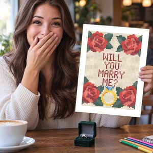 Puede incluir: Un patr&oacute;n de punto de cruz rectangular sobre tela blanca que presenta cuatro rosas rojas con hojas verdes en las esquinas y el texto WILL YOU MARRY ME? en letras rojo oscuro, con un icono de anillo de compromiso amarillo en el centro inferior.