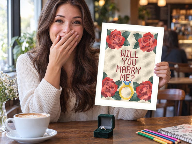 Puede incluir: Un patr&oacute;n de punto de cruz rectangular sobre tela blanca que presenta cuatro rosas rojas con hojas verdes en las esquinas y el texto WILL YOU MARRY ME? en letras rojo oscuro, con un icono de anillo de compromiso amarillo en el centro inferior.