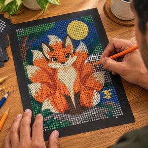May include: A pixel art style colouring page featuring a multi-tailed fox with orange and white fur, large eyes, and a yellow moon in a dark blue night sky. The image is composed of a grid of small squares, with coloured pencils visible nearby.
