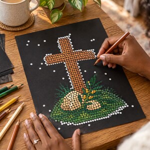 K&ouml;nnte beinhalten: Ein schwarzes Blatt Papier mit einer Punkt-Illustration eines braunen Kreuzes, umgeben von gr&uuml;nem Blattwerk und kleinen wei&szlig;en Punkten. Das Kunstwerk ist in Pointillismus-Technik erstellt und mehrere Buntstifte liegen auf dem Tisch bereit.
