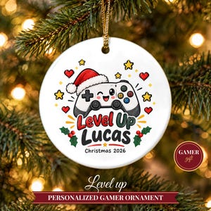 Peut inclure: Une décoration ronde en céramique blanche illustrant une manette de jeu vidéo de dessin animé portant un bonnet de Père Noël. Le texte indique Level Up Lucas Christmas 2026, entouré de cœurs, d'étoiles et de houx pixelisés.