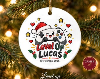 May include: A round white ceramic ornament featuring a cartoon video game controller wearing a Santa hat. The text reads Level Up Lucas Christmas 2026, surrounded by pixelated hearts, stars, and holly leaves, hanging from a gold cord.