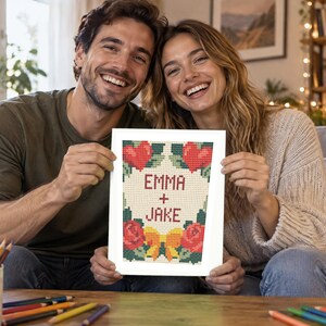 Op de afbeelding: Een rechthoekig kruissteekpatroon gedrukt op wit papier met de tekst EMMA + JAKE in het rood. Het ontwerp bevat twee rode harten bovenaan, twee rode rozen onderaan, groene bladeren en een gele strik gecentreerd aan de onderkant.
