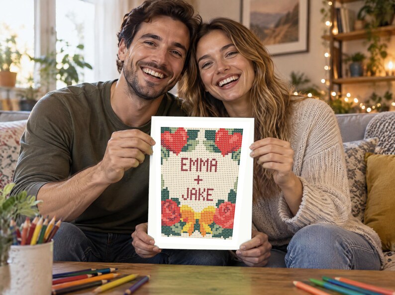 Op de afbeelding: Een rechthoekig kruissteekpatroon gedrukt op wit papier met de tekst EMMA + JAKE in het rood. Het ontwerp bevat twee rode harten bovenaan, twee rode rozen onderaan, groene bladeren en een gele strik gecentreerd aan de onderkant.
