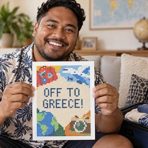 May include: A rectangular pixel art print featuring the text OFF TO GREECE! in dark blue letters. The design includes a red suitcase, a blue airplane, a green and blue globe, and a brown camera, all rendered in a colorful grid-based mosaic style.
