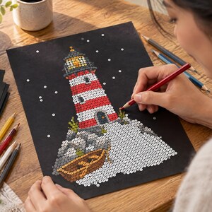 May include: A black craft sheet featuring a sequin art project of a red and white striped lighthouse on a rocky base with a small brown boat. The design includes a yellow light at the top, grey rocks, and white accents, with scattered sequins.