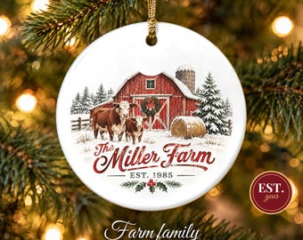 May include: A round white ceramic Christmas ornament featuring a red barn, a silo, a snow-covered landscape, and two brown and white cows. The text reads The Miller Farm EST. 1985, with a festive wreath on the barn and holly berries below.
