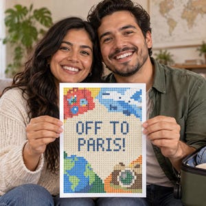 May include: A rectangular diamond painting canvas featuring the text OFF TO PARIS! in blue block letters. The design includes a red suitcase, a blue airplane, a green and blue globe, and a brown camera, all rendered in a pixelated grid pattern.