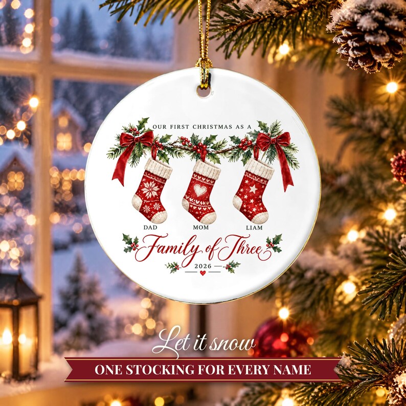 May include: A round white ceramic Christmas ornament featuring three red and white patterned stockings hanging from a holly branch with red bows. Text reads: Our First Christmas as a Family of Three, Dad, Mom, Liam, and 2026.