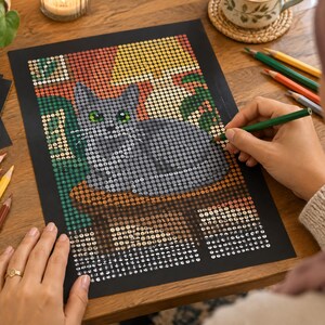 Pu&ograve; includere: Un progetto artistico rettangolare su carta nera con una griglia di piccoli adesivi circolari che formano un gatto grigio con occhi verdi. Il gatto &egrave; seduto su una superficie marrone davanti a uno sfondo di forme gialle e verdi.