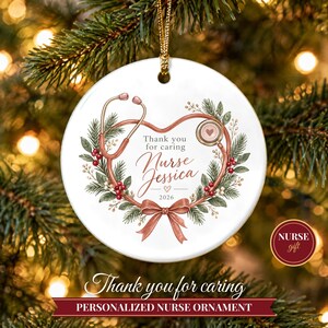 Peut inclure: Une décoration ronde en céramique blanche ornée d'un stéthoscope en forme de cœur, entouré d'une couronne de branches de pin et de baies rouges. Le texte indique Thank you for caring, Nurse Jessica, et 2026, avec un ruban rose.