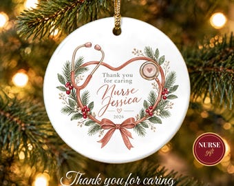 May include: A round white ceramic ornament featuring a stethoscope shaped into a heart, surrounded by a wreath of pine branches and red berries. The text reads Thank you for caring, Nurse Jessica, and 2026, accented with a pink ribbon.