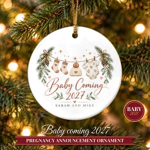 May include: A round white ceramic ornament featuring a clothesline illustration with two socks, a knit hat, and a baby onesie. The text reads Baby Coming 2027 and Sarah and Mike. It is decorated with pine branches, pinecones, and gold stars.